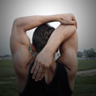 A teenage boy stands in a field doing an overhead tricep stretch.