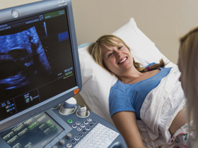 A smiling blond woman in a blue shirt lies on an exam table. Dr. Howley looks at a screen on a computer-like machine showing fetal ultrasound results. 
