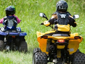 Three kids ride on two ATVs through a grassy field.