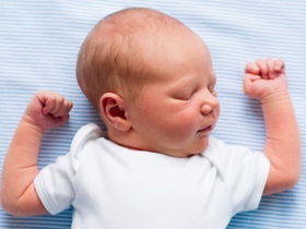 A baby sleeps on its back in a crib with hands up near head.