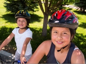 Two girls wearing helmets stand on their bikes in a park.